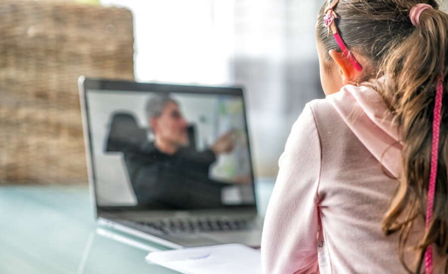 Girl following school lesson on her laptop with male teacher speaking